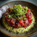 A vibrant serving of tuna tartare, featuring diced red tuna over a creamy green avocado base, topped with sesame seeds and curled scallions.