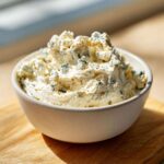 A close-up of a bowl filled with chunky, creamy blue cheese dip, highlighted by bright sunlight.