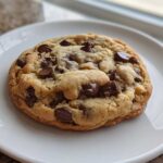A single, thick, and chewy Chocolate Chip Cookie loaded with melted chocolate chips, resting on a white plate.