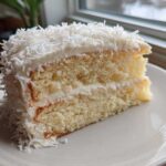 A generous slice of moist coconut cake recipe layered with white frosting and covered in shredded coconut.