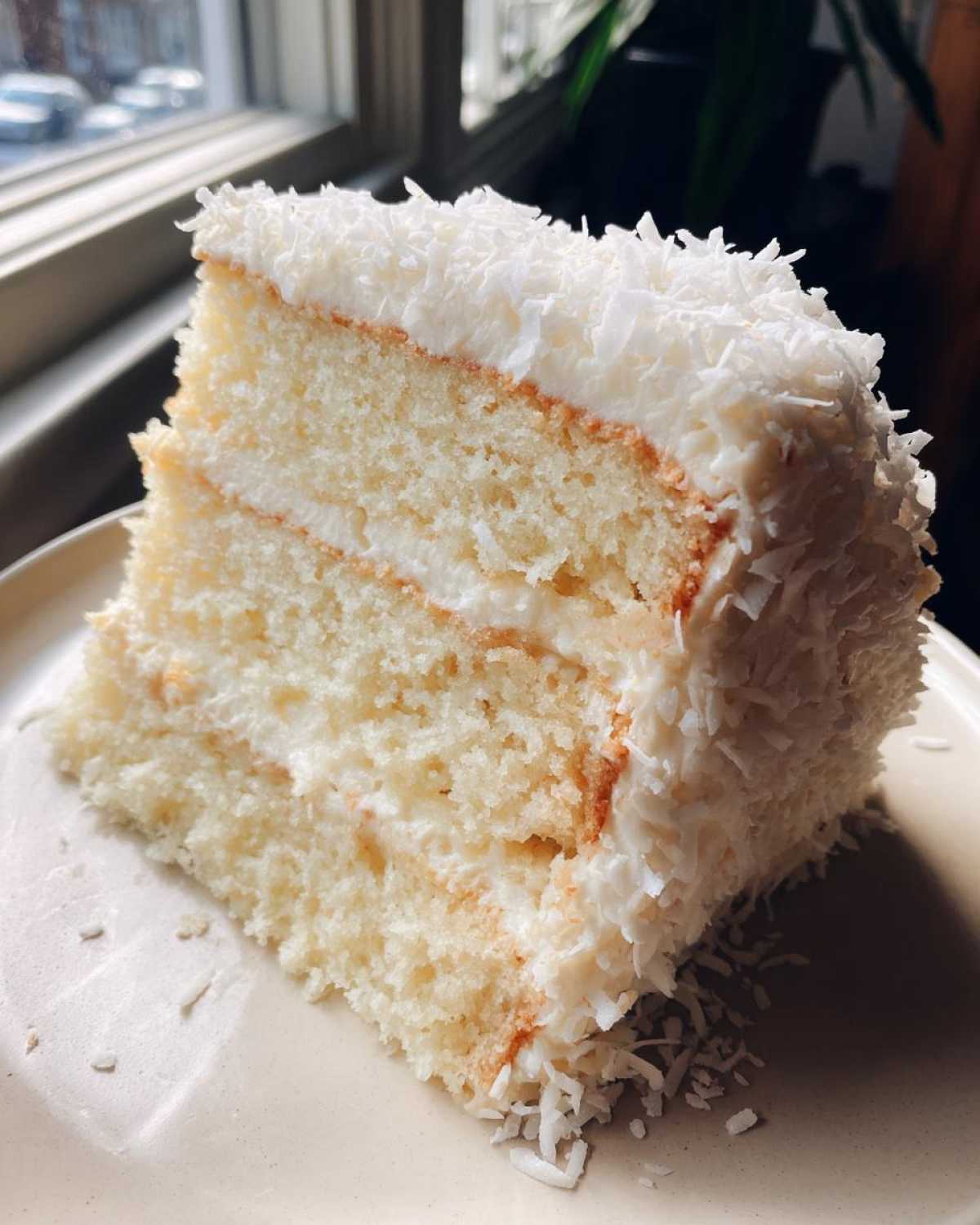 Close-up of a moist, three-layer slice of coconut cake recipe covered in white frosting and shredded coconut.