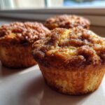 Three golden brown coffee cake muffins featuring a rich, sugary cinnamon streusel topping, sitting on a light surface near a window.