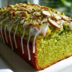A vibrant green pistachio bread loaf drizzled with white glaze and topped with sliced almonds and chopped pistachios.