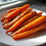 A close-up of glossy, bright orange roasted carrots seasoned with herbs on a white plate.