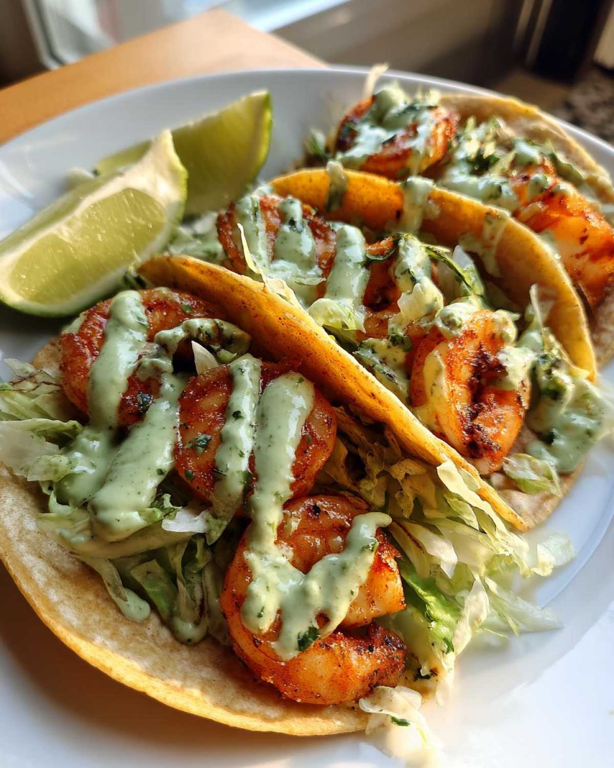 Three delicious Shrimp Tacos filled with seasoned shrimp, shredded lettuce, and drizzled with a creamy green sauce, served with a lime wedge.