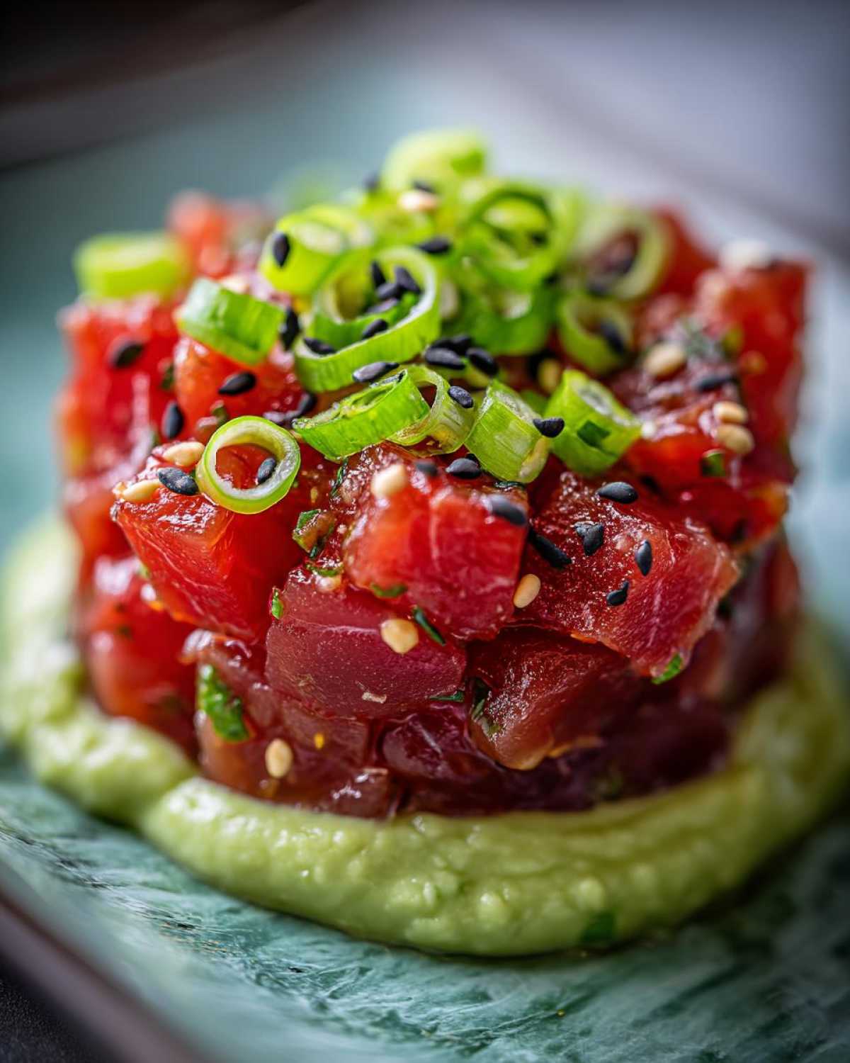 Close-up of vibrant red tuna tartare stacked high on a creamy avocado puree, topped with green onions and black sesame seeds.