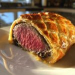 Close-up of a slice of Authentic Beef Wellington showing medium-rare beef inside golden, lattice-patterned puff pastry.