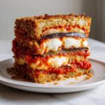 A tall, layered slice of rich Eggplant Parmesan Recipe Authentic, showing breaded eggplant, melted mozzarella, and bright tomato sauce.