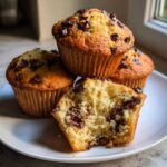 Close-up of three chocolate chip muffins, one broken open showing a moist, fluffy interior packed with chocolate chips.