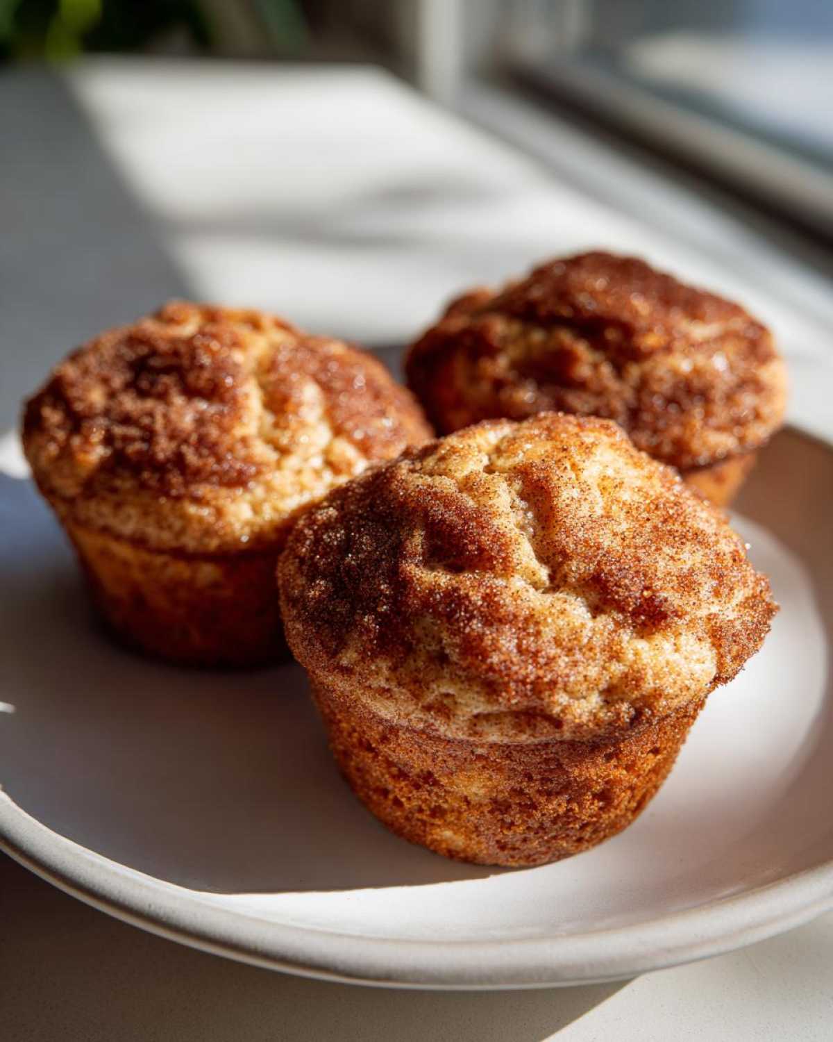 Three freshly baked Banana Cinnamon Muffins with a crunchy cinnamon sugar topping resting on a light gray plate.
