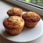 Three freshly baked Banana Cinnamon Muffins with a sugary cinnamon crust sitting on a white plate near a window.