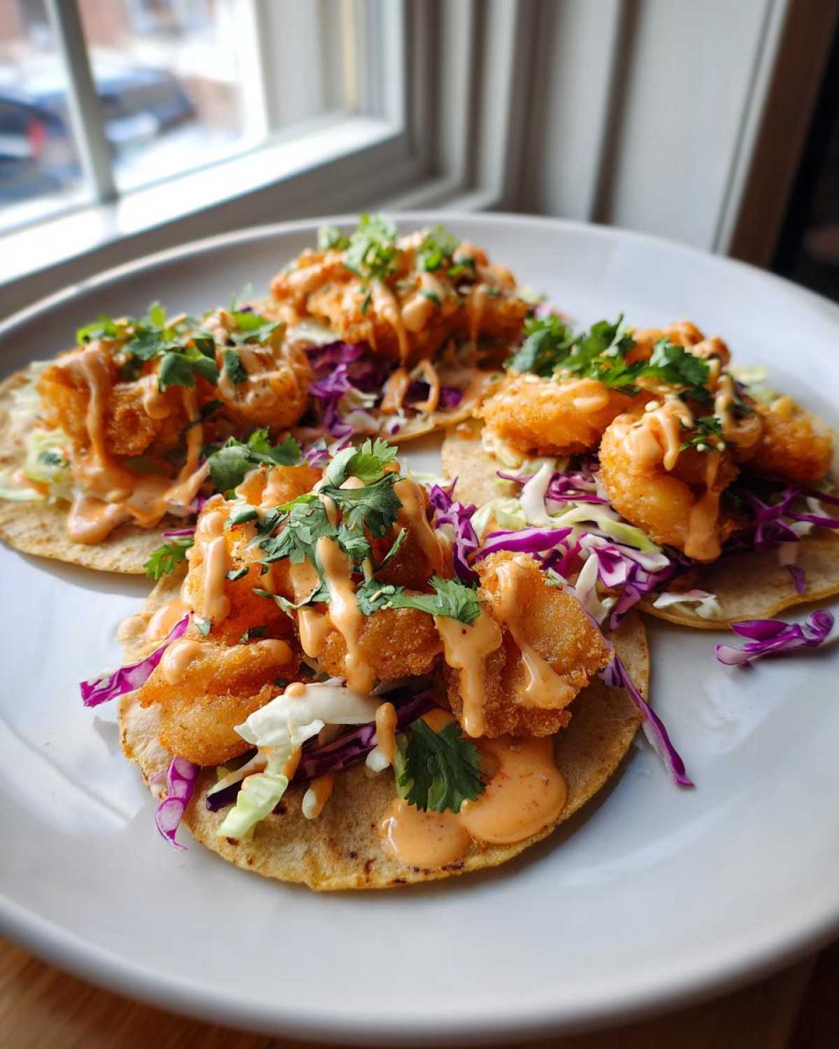 Four delicious Bang Bang Shrimp Tacos served on a white plate, topped with creamy sauce and cilantro.