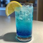 Close-up of a tall glass containing a vibrant Blue Drinks Aesthetic Cocktail filled with ice and garnished with a fresh lemon slice.