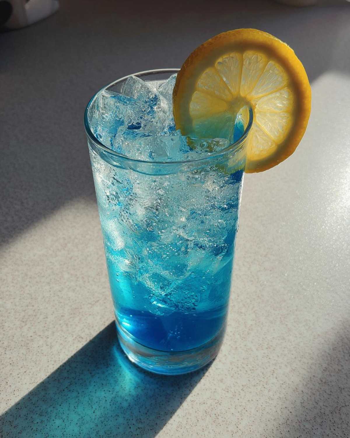 A tall glass filled with a bright Blue Drinks Aesthetic Cocktail, ice, and garnished with a fresh lemon wheel.