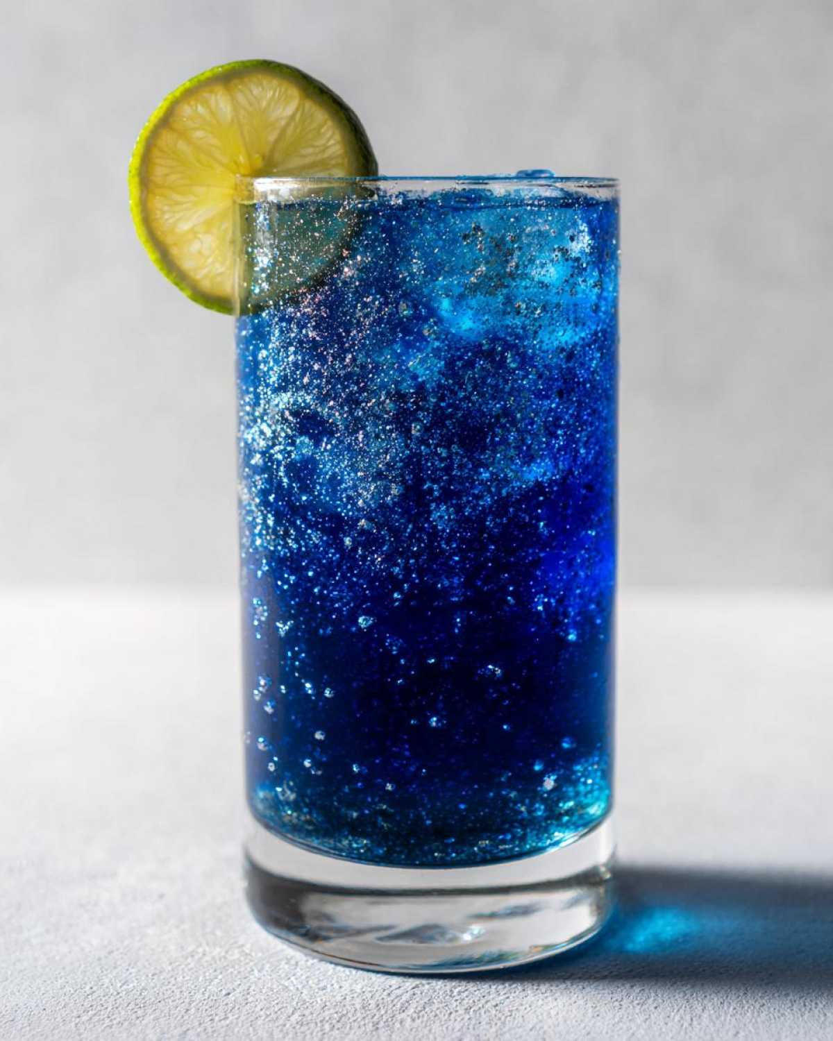 A tall glass filled with a vibrant, deep blue drinks aesthetic mocktail featuring edible glitter and a fresh lime wheel garnish.