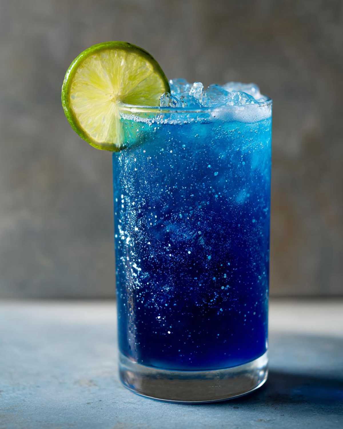 A tall glass filled with a vibrant, shimmering Blue Drinks Aesthetic Mocktail, topped with crushed ice and garnished with a fresh lime wheel.