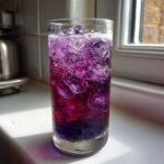 A tall glass filled with a vibrant, layered purple and red Blueberry Jam Gin Spritz, covered in condensation and ice.