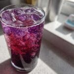 Close-up of a tall glass filled with a vibrant, bubbly Blueberry Jam Gin Spritz, showing deep purple and red hues over ice cubes.