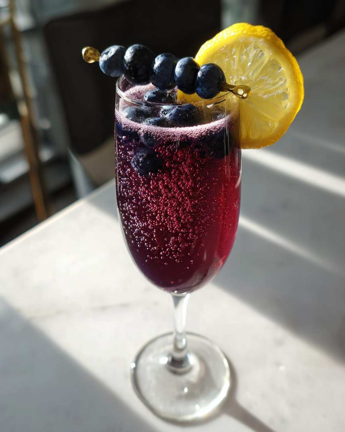 A vibrant, bubbly Blueberry Lemosas Brunch Cocktail garnished with fresh blueberries on a skewer and a lemon slice.
