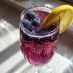 A vibrant Blueberry Lemosas Brunch Cocktail, bubbly and purple, garnished with fresh blueberries and a lemon slice.