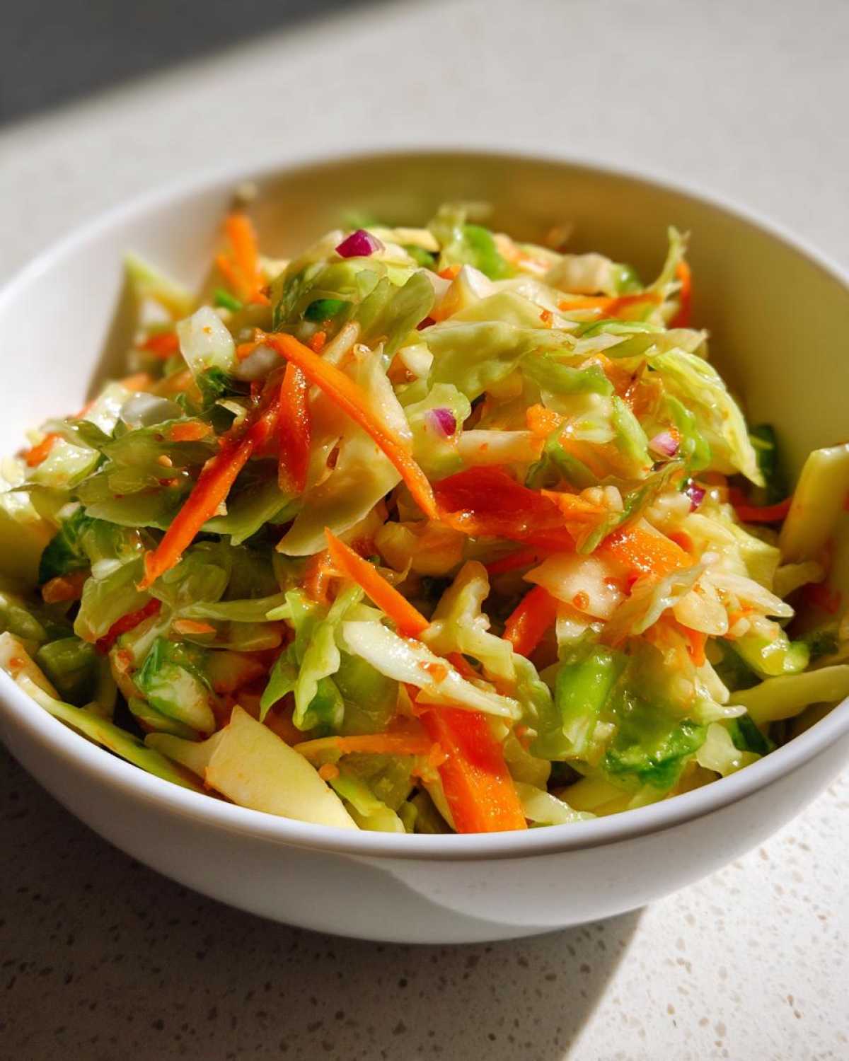 A close-up of vibrant Cabbage and Apple Crispy Slaw featuring shredded cabbage, carrots, and apple chunks in a white bowl.
