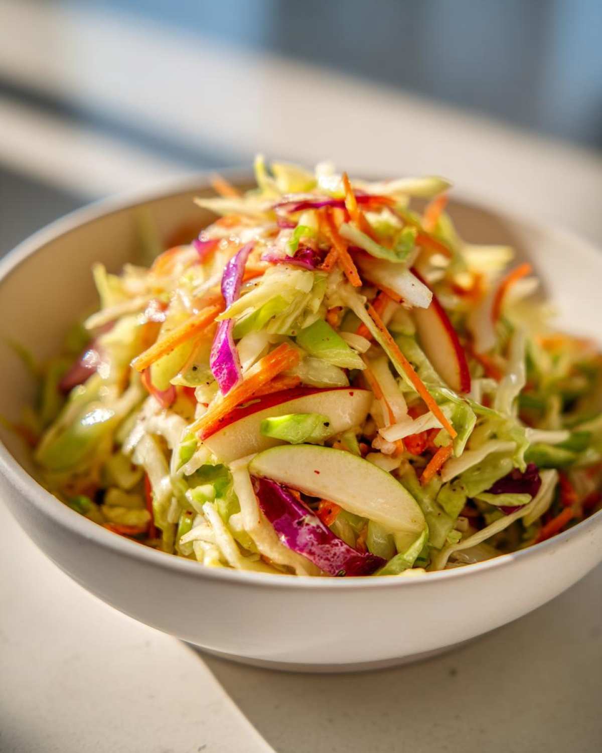 A close-up of vibrant Cabbage and Apple Crispy Slaw featuring shredded cabbage, carrots, red cabbage, and apple slices in a white bowl.