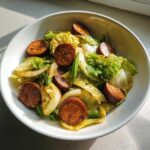 A close-up of a serving of Cabbage and Sausage Skillet in a white bowl, featuring browned sausage slices and wilted cabbage.