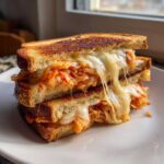 A close-up of a Cabbage Kimchi Grilled Cheese sandwich cut in half and stacked, showing melted, stretching cheese and bright red kimchi filling.
