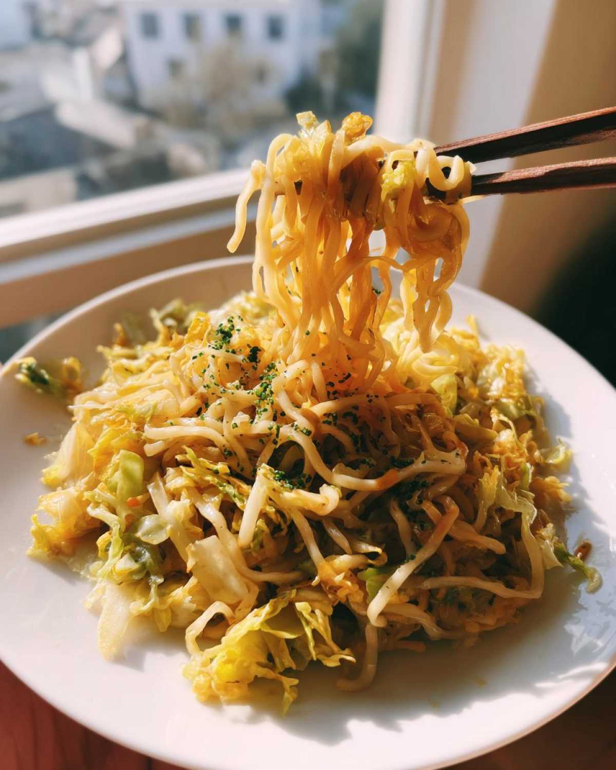 A close-up of someone lifting noodles and cabbage from a plate of Cabbage Noodle Stir Fry 10 Mins using dark wooden chopsticks.