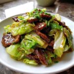 A close-up of glossy Cabbage and Pork Stir Fry Ginger served on a white plate, topped with fresh chives.