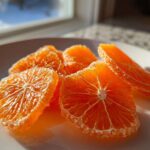 Close-up of several bright orange candied orange slices coated in sugar resting on a white plate near a window.