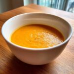 A close-up of a white bowl filled with vibrant orange carrot ginger soup, seasoned with black pepper.