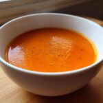 A close-up of a white bowl filled with bright orange carrot ginger soup, lightly seasoned with black pepper.