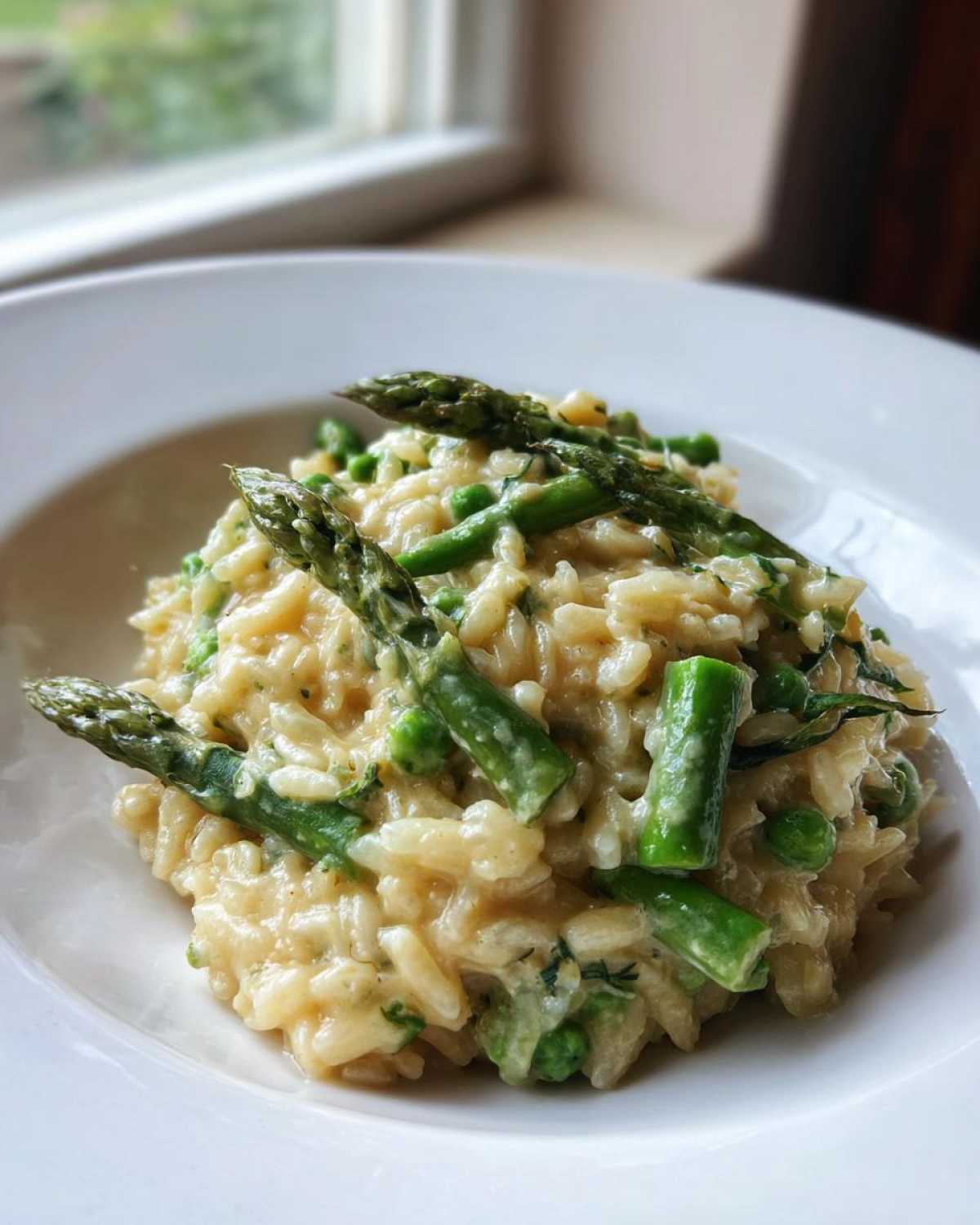 A close-up of creamy Cheesy Asparagus Orzotto topped with bright green asparagus spears and peas in a white bowl.