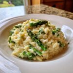 A close-up of creamy Cheesy Asparagus Orzotto, garnished with bright green asparagus tips, served in a white bowl.