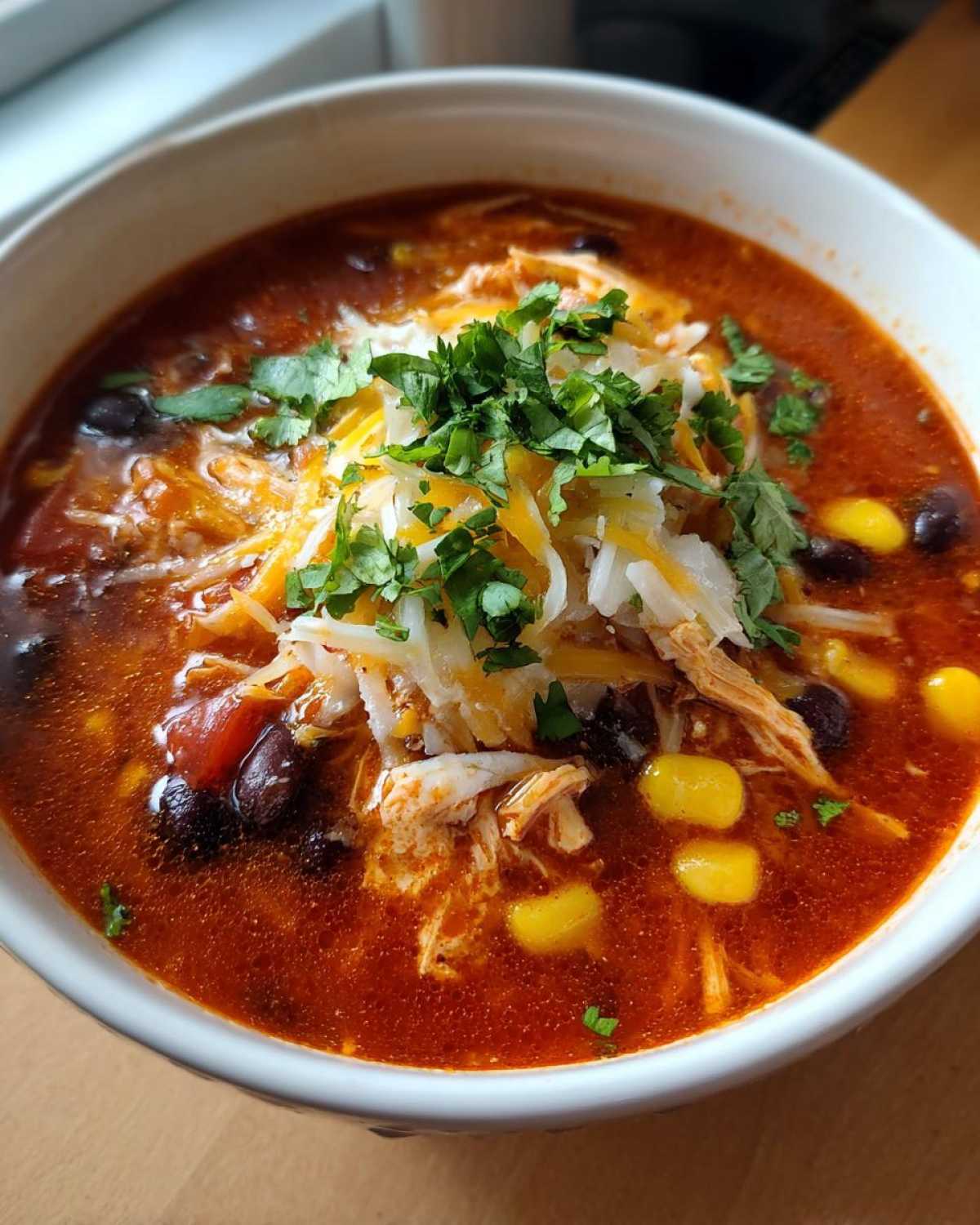 Close-up of a bowl of rich, red chicken enchilada soup topped with shredded cheese, sour cream, and fresh cilantro.