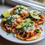 Three delicious Chicken Tinga Tacos Authentic served on a white plate, topped with crumbled cheese, avocado slices, and cilantro.