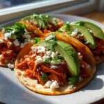 Four assembled Chicken Tinga Tacos on a plate, topped with crumbled white cheese, cilantro, and fresh avocado slices.