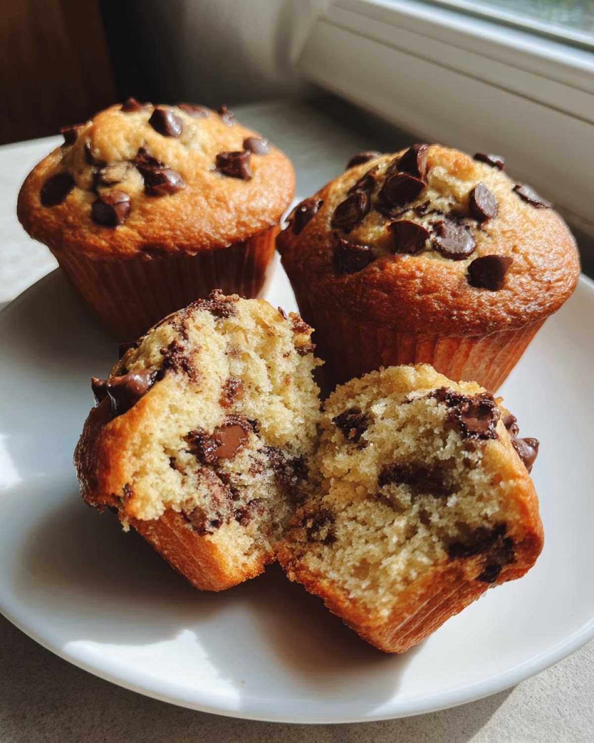 Three golden brown chocolate chip muffins on a white plate, one split open showing the moist interior.