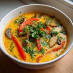 A close-up of a bowl of vibrant yellow coconut curry soup, topped with sliced red peppers, mushrooms, and fresh cilantro.