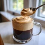 A spoonful of thick Cookie Butter Cold Foam is being added to a glass mug of dark iced coffee.