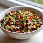 Close-up of a white bowl filled with vibrant Cowboy Caviar, featuring black beans, corn, red onion, and peppers.