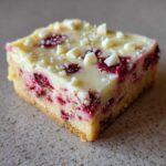 A single square of cranberry bliss bars showing the shortbread base, bright red cranberry filling, and white chocolate frosting with chips.