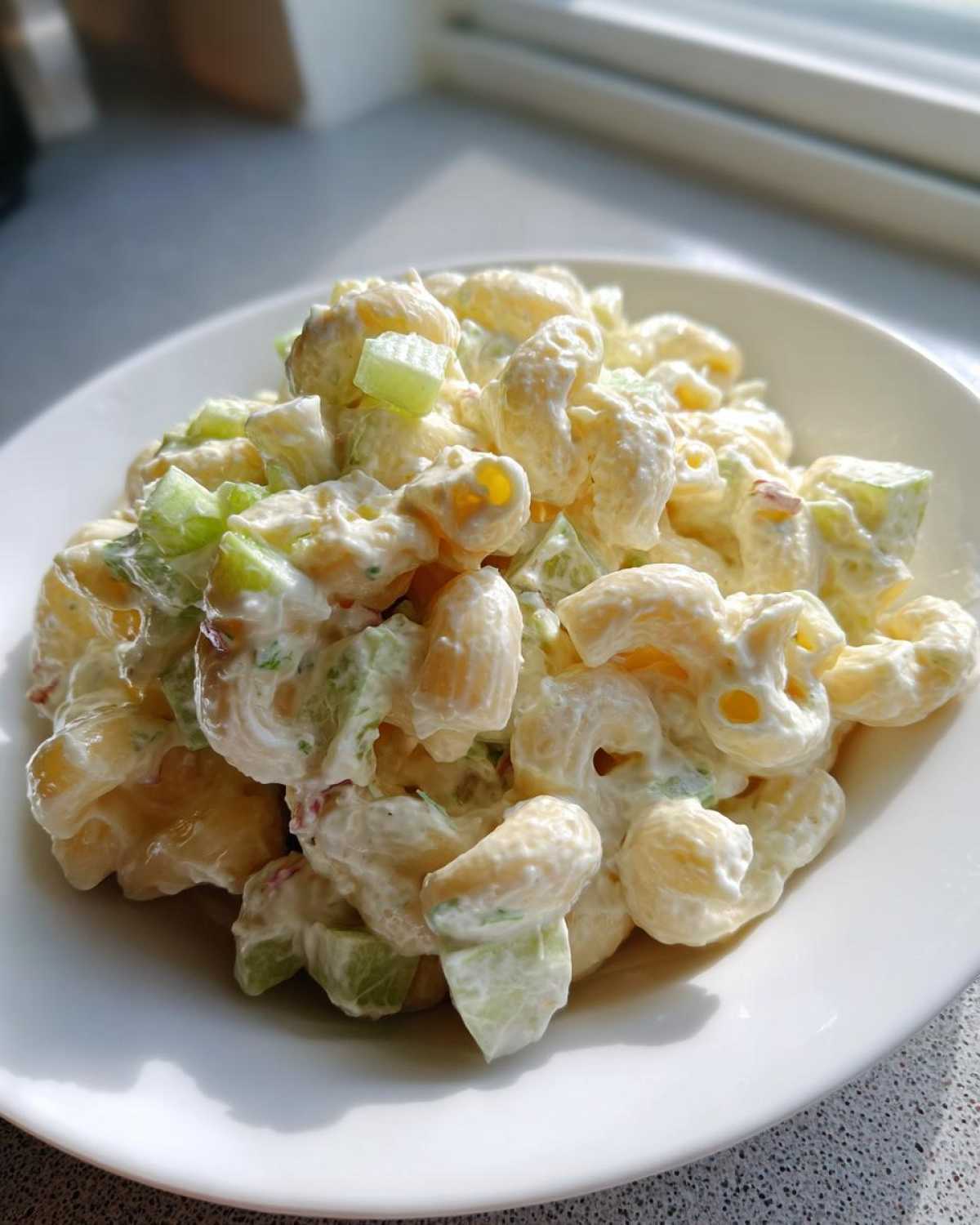 A close-up of creamy Amish macaroni salad mixed with chopped celery served in a white bowl near a window.