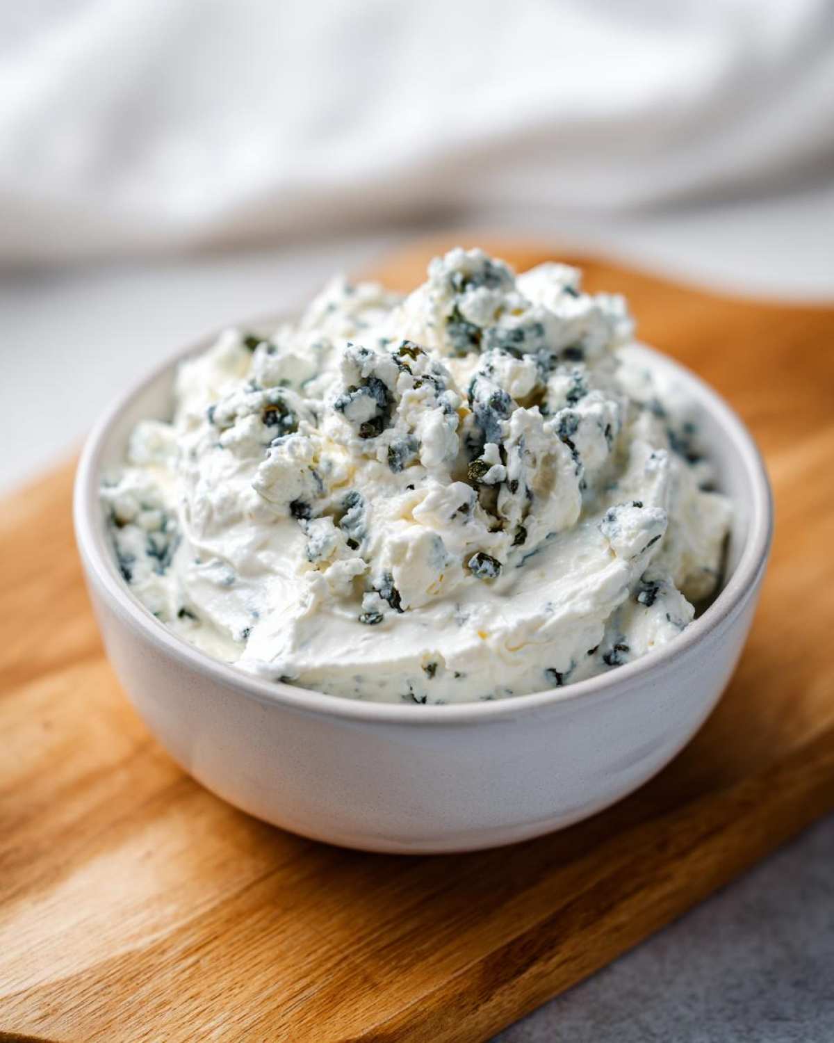 Close-up of a white bowl filled with chunky, creamy blue cheese dip, resting on a wooden cutting board.