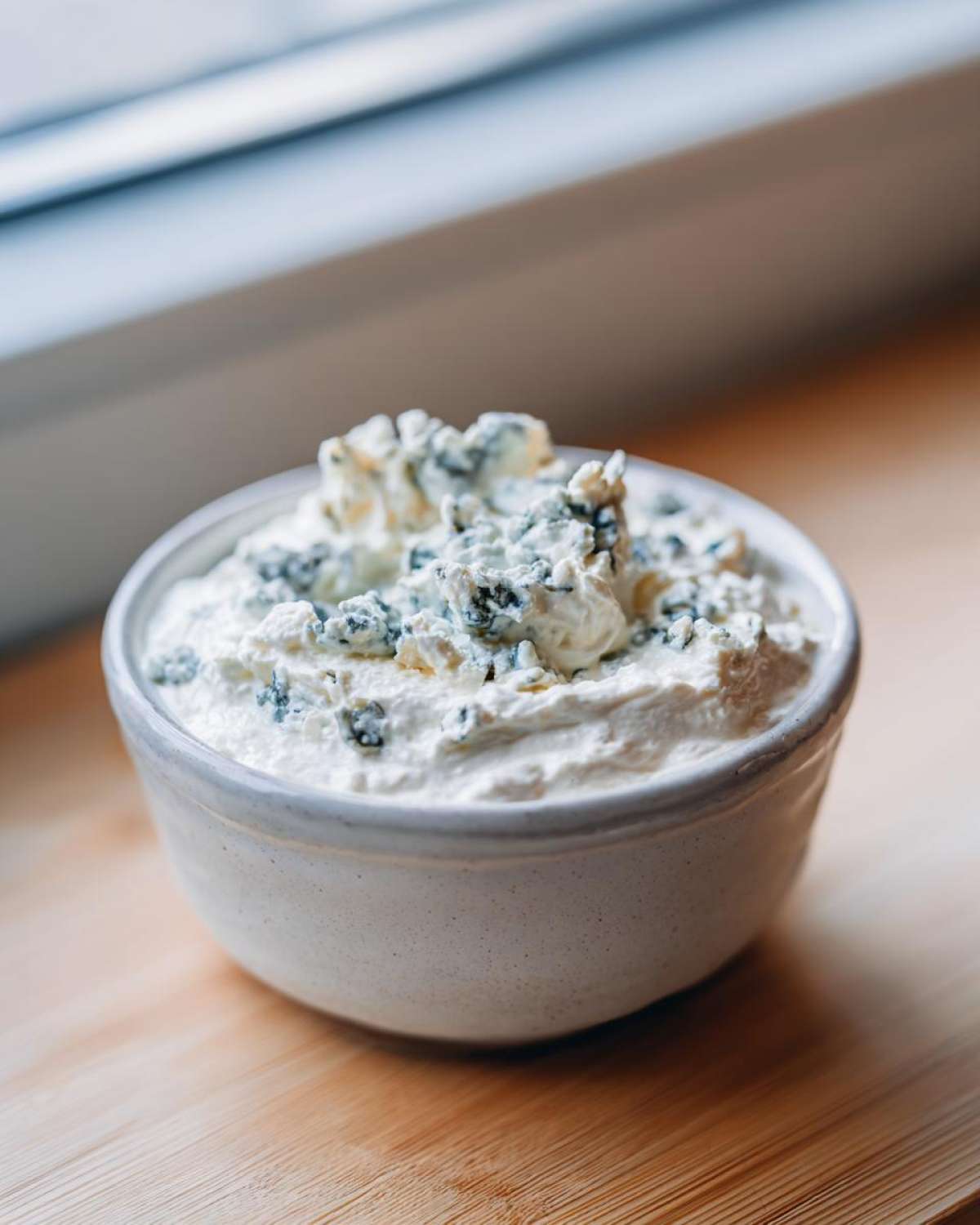 A small white bowl filled with creamy blue cheese dip, topped generously with blue cheese crumbles.
