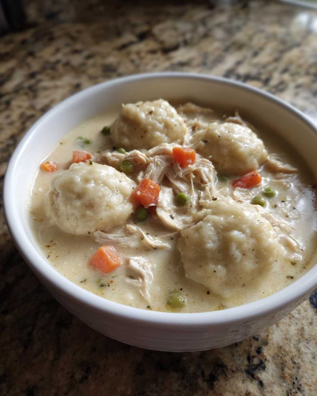 Close-up of a white bowl filled with creamy chicken and dumpling soup, featuring shredded chicken, carrots, and peas.