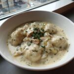 A close-up of a white bowl filled with creamy chicken and dumplings, garnished with fresh parsley.