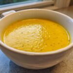 Close-up of a white bowl filled with bright yellow, creamy corn soup, lightly seasoned with pepper.