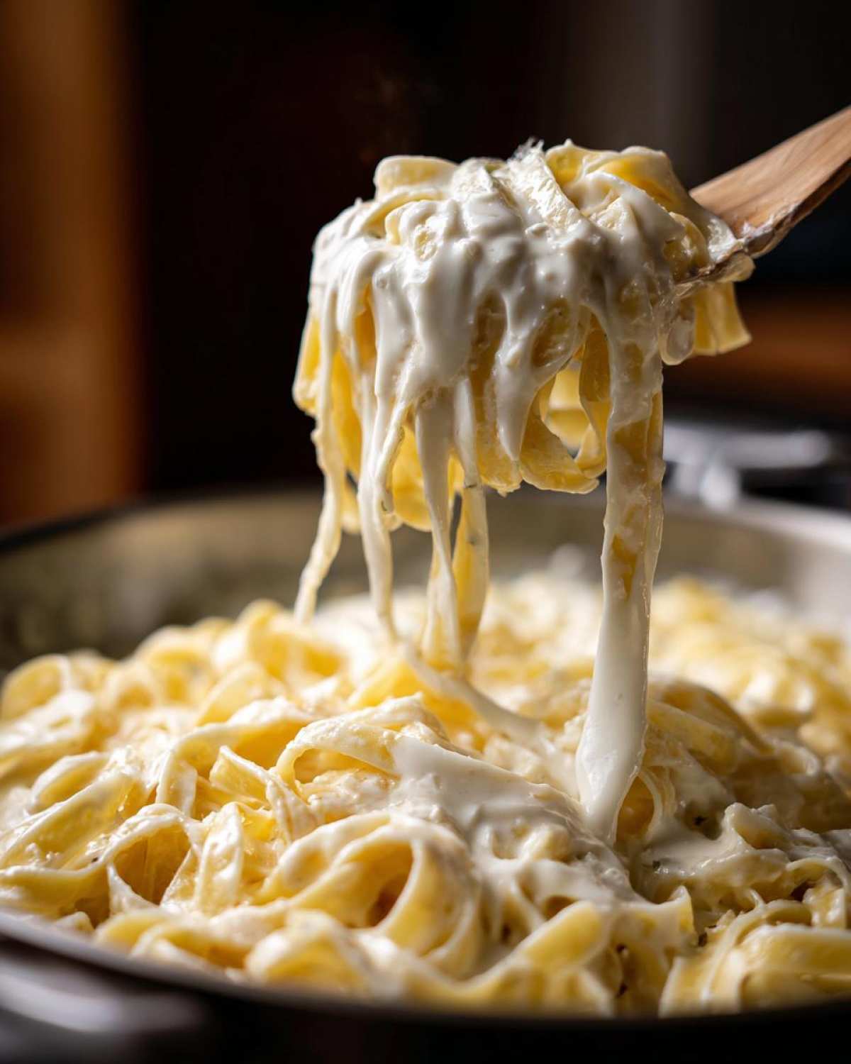 A wooden spoon lifts fettuccine noodles coated in thick, creamy cottage cheese alfredo sauce from a pan.